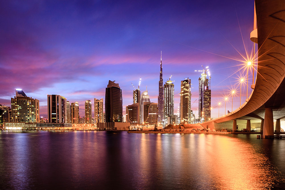 Vista panorâmica do skyline moderno de Dubai ao crepúsculo, destacando o Burj Khalifa ilumina-se ao centro, com uma ponte iluminada em primeiro plano refletindo luzes douradas na água.