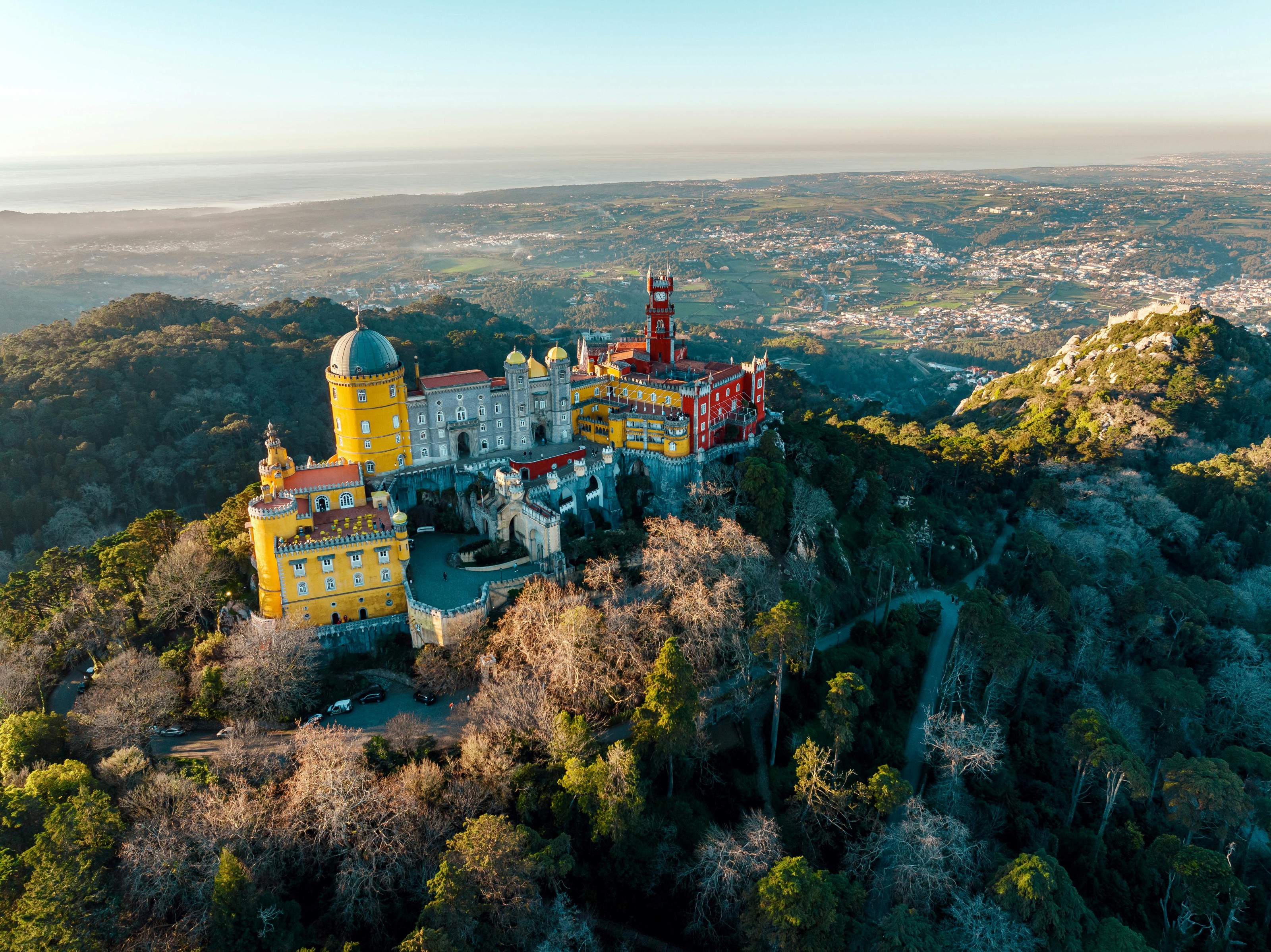 Vista aérea do Palácio da Pena em Sintra, monumento romântico colorido entre florestas verdejantes e paisagem atlântica, Património Mundial da UNESCO.