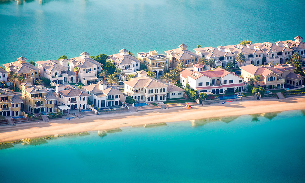 Vista aérea de vilas residenciais de luxo à beira-mar na ilha de Palm Jumeirah, com praia de areia dourada e águas azul-turquesa.